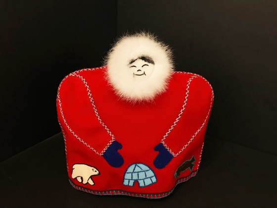 This tea cozy is in the shape of a smiling person wearing a red parka with white fur around their face. White stitching outlines the sleeves, where blue mittens poke out from. A polar bear, an igloo, and a bison are stitched onto the parka along the bottom. 