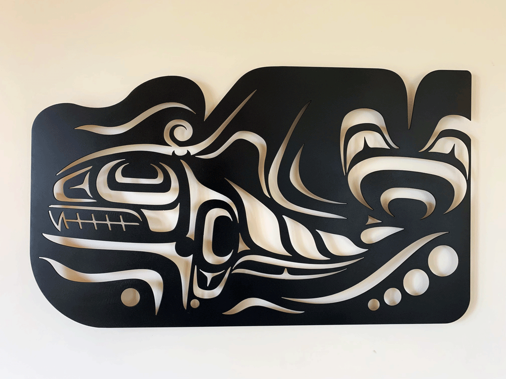 A black metal Coastal Salish wall sculpture on a white background. Crescents, u-shapes and trigons carved out of a metal sheet create the form of an imposing Orca whale with a tall dorsal fin and strong tail. The top edge of the metal sheet has also been carved to create the impression of water moving in response to the powerful motion of the whale.