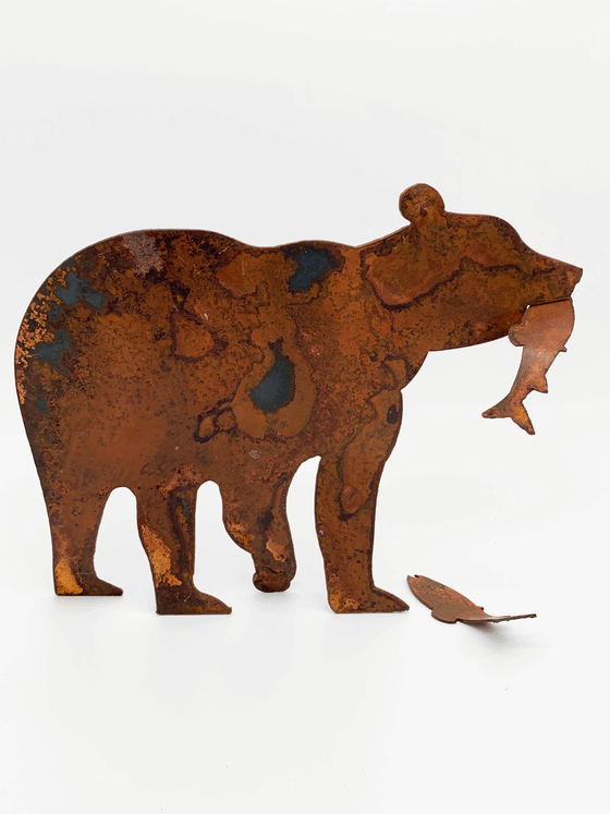 A bear silhouette standing on four legs, holding a detachable salmon in its mouth, cut from warm, weathered steel. At its feet lies a second salmon. The unique dappling of the oxidized steel makes each piece one-of-a-kind.