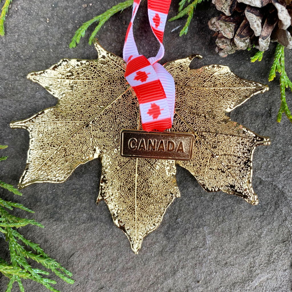 A large gold coated maple leaf sits on a stone background. A ribbon patterned with Canadian flags is tied to the stem end of the leaf. On the ribbon is a small gold charm that says “Canada”. The gold has a bright finish. Around the picture are decorative evergreen leaves and a pine cone.