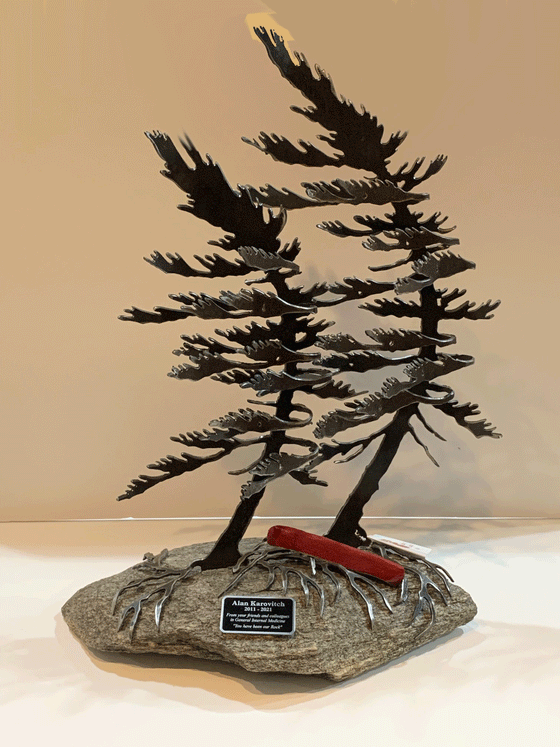 Steel windswept pine trees fitted onto a piece Canadian stone. A red canoe rests on the roots underneath  the trees. An engraved plaque is fixed on the front of the stone.
