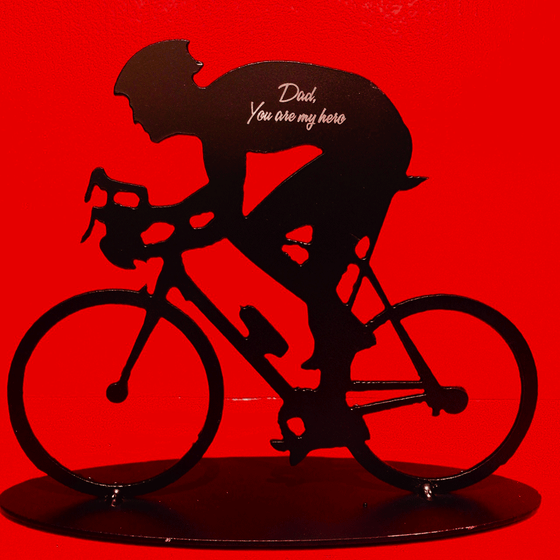 A metal sculpture of a male cyclist. The sculpture has been painted matte black. Direct engraving of the words “Dad, You are my hero” onto cyclist’s back makes the silver of the underlying metal shine against the black paint.