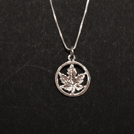 A small sterling silver maple leaf on a silver chain sits on a black background. The maple leaf is enclosed by a silver circle. The chain attaches to the top of the circle.