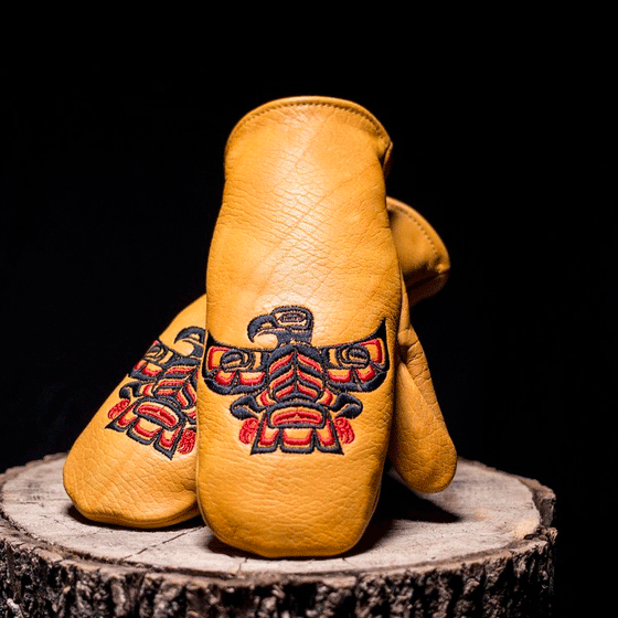 Yellow mittens with Haida Eagle on the part where back of hand goes. Mittens are sitting on a tree stump.