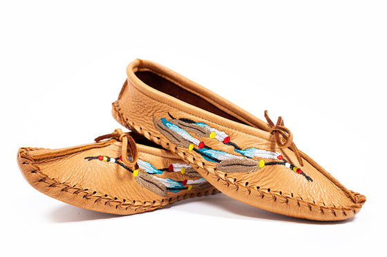 Tan brown moccasins with embroidered beads and feathers of a dream catcher on the side with a lace to adjust it. Moccasins are sitting balanced on top of each other with a white background. 