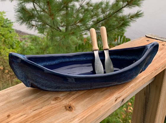 A stoneware canoe sits on a weathered wooden railing. Two spreaders shaped like paddles rest against the gunnel. The lower half of the canoe is deep blue, with a contrasting rim in mottled lighter blue. A peaceful background of trees and lake complete the picture.