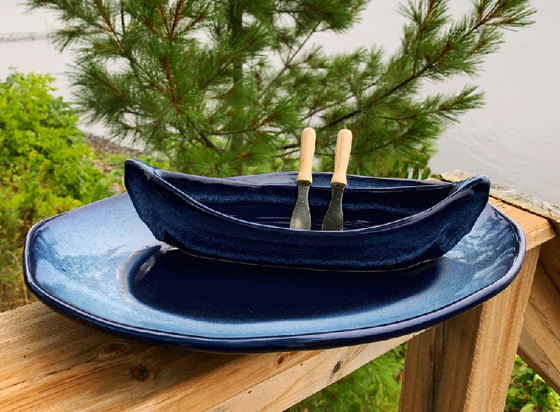 A stoneware canoe sits on a matching stoneware serving plate atop a weathered wooden railing. Two spreaders shaped like paddles rest against the gunnel. The base of both the canoe and platter are deep blue, with contrasting rims in mottled lighter blue. A peaceful background of trees and lake complete the picture.
