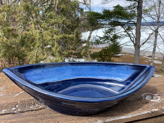 Bottom half of bowl is off-white. Top half of bowl is blue with white accents. Bowl is oval shaped with points at two opposite ends Bowl is on a piece of wood with trees and a lake in the background.