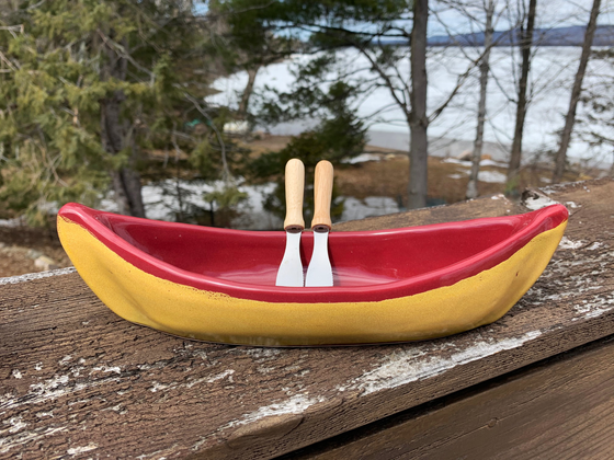 A stoneware canoe sits on a weathered wooden railing. Two spreaders shaped like paddles rest against the gunnel. The lower half of the canoe is golden yellow, while the rim and interior are deep red. A peaceful background of trees and lake complete the picture.