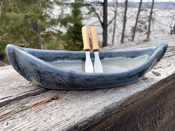 A stoneware canoe sits on a weathered wooden railing. Two spreaders shaped like paddles rest against the gunnel. The lower half of the canoe is grey, with a contrasting rim in mottled blue. A peaceful background of trees and lake complete the picture.