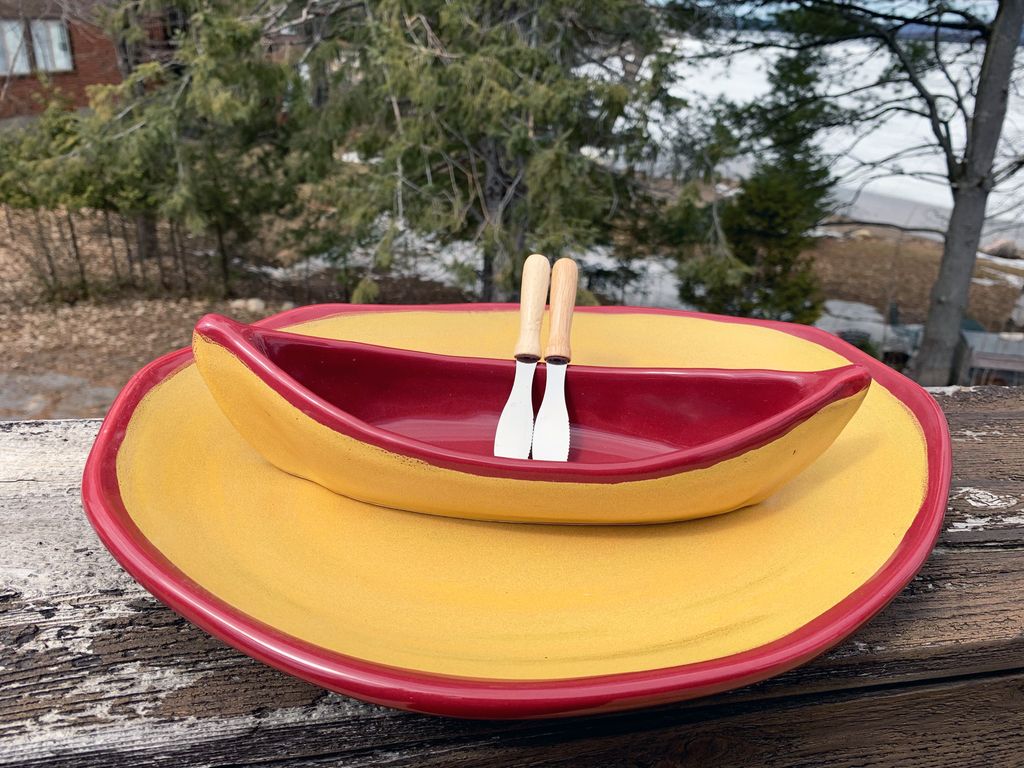 A stoneware canoe sits on a matching stoneware serving plate atop a weathered wooden railing. Two spreaders shaped like paddles rest against the gunnel. The base of both the canoe and platter are golden yellow, with contrasting rims in deep red. A peaceful background of trees and lake complete the picture.