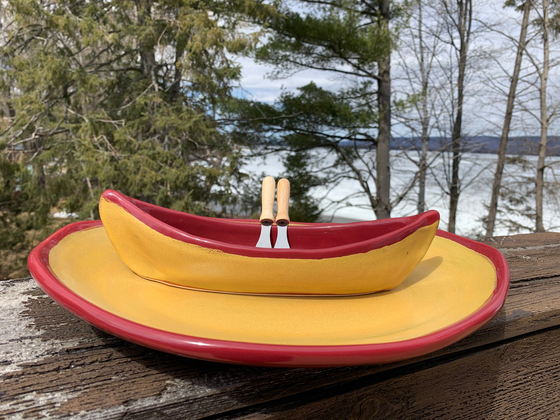 A stoneware canoe sits on a matching stoneware serving plate atop a weathered wooden railing. Two spreaders shaped like paddles rest against the gunnel. The base of both the canoe and platter are golden yellow, with contrasting rims in deep red. A peaceful background of trees and lake complete the picture.