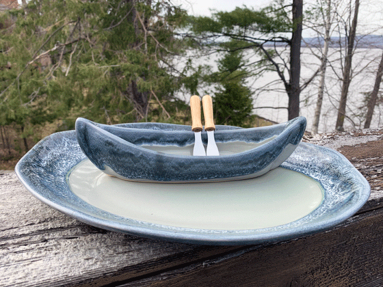 A stoneware canoe sits on a matching stoneware serving plate atop a weathered wooden railing. Two spreaders shaped like paddles rest against the gunnel. The base of both the canoe and platter are greenish-grey, with contrasting rims in mottled blue and white. A peaceful background of trees and lake complete the picture.
