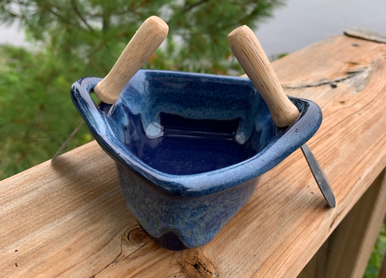 A stoneware rowboat sits on a weathered wooden railing. Two spreaders shaped like paddles sit in the oarlocks. The base is deep blue, with a contrasting rim in mottled lighter blue and white. A peaceful background of trees and lake complete the picture.