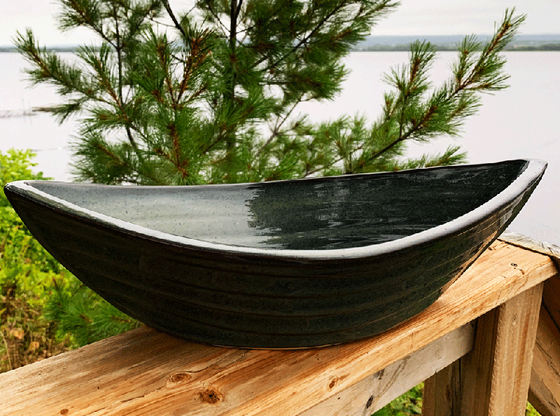 Sage green bowl. Bowl is oval shaped with points at two opposite ends. Bowl is on a piece of wood with trees and a lake in the background.