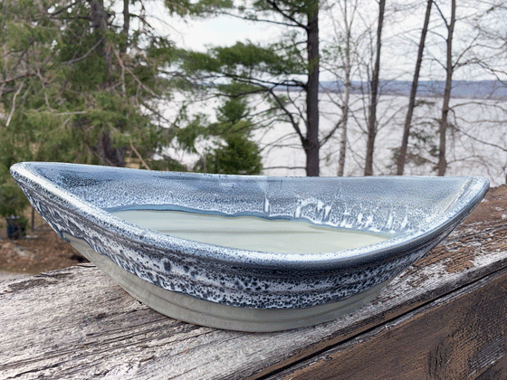 Bottom half of bowl is blue-grey. Top half of bowl is blue with white accents. Bowl is oval shaped with points at two opposite ends. Bowl is on a piece of wood with trees and a lake in the background.