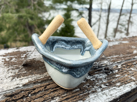 A stoneware rowboat sits on a weathered wooden railing. Two spreaders shaped like paddles sit in the oarlocks. The base is greenish-grey, with a contrasting rim in mottled blue, black, and white. A peaceful background of trees and lake complete the picture.