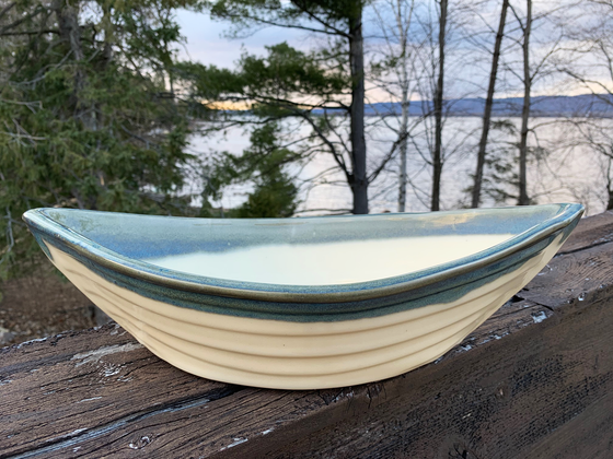 Bowl is off-white with a layer of blue colour around the top edge. Bowl is oval shaped with points at two opposite ends. Bowl is on a piece of wood with trees and a lake in the background.