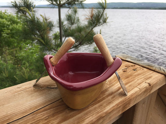A stoneware rowboat sits on a weathered wooden railing. Two spreaders shaped like paddles sit in the oarlocks. The base is golden yellow, with a contrasting rim and interior is deep red. A peaceful background of trees and lake complete the picture.