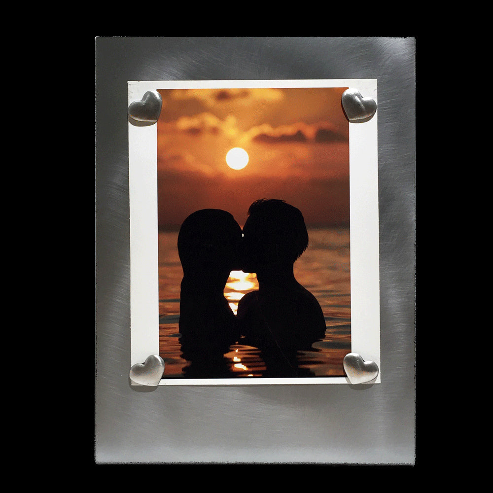 A vertical rectangular metal picture frame with four heart shaped pewter magnets holding a picture of two people swimming at sunset.