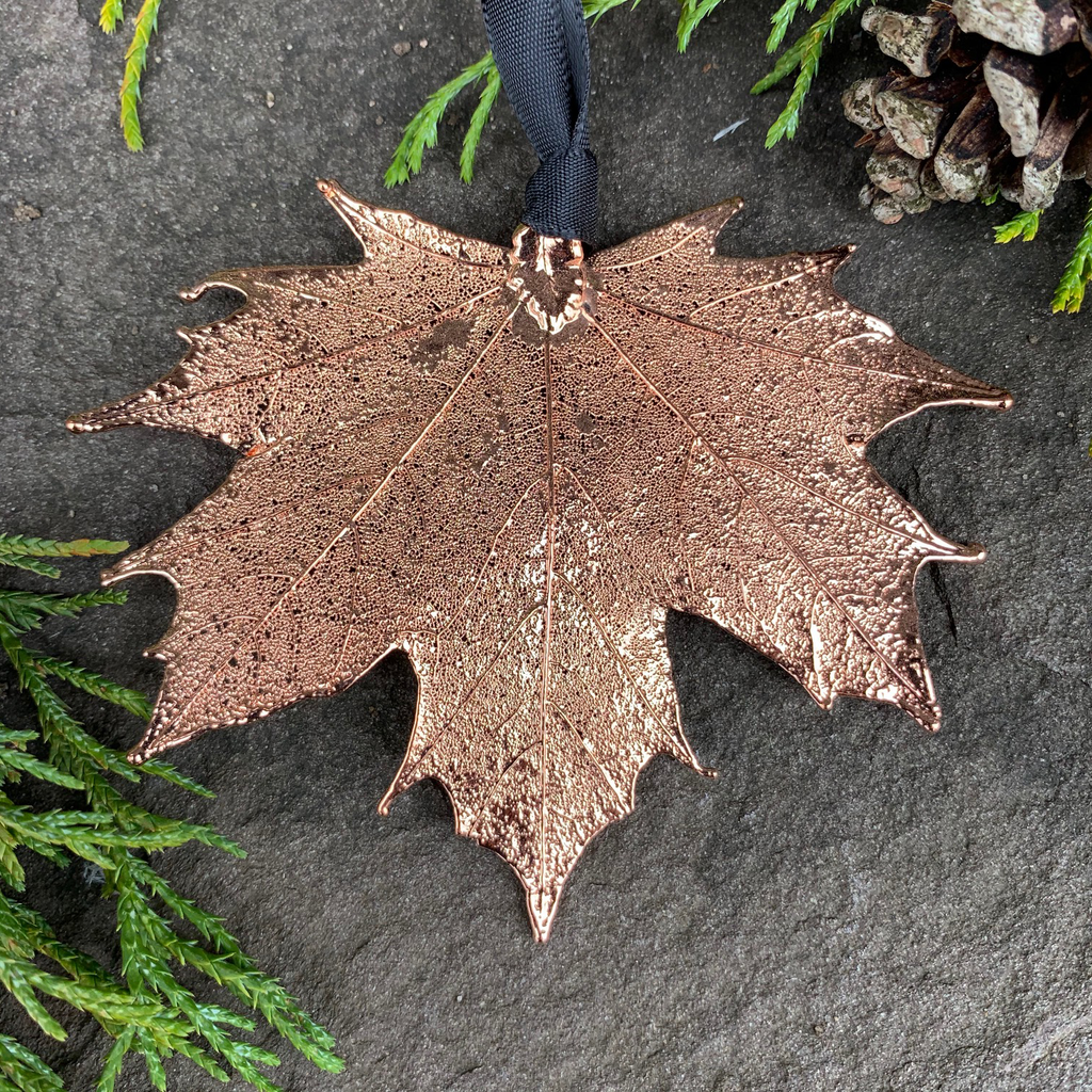 A large copper coated maple leaf sits on a stone background. A black ribbon is tied to the stem end of the leaf. The copper has a bright finish. Around the picture are decorative evergreen leaves and a pinecone.
