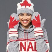 A smiling woman wearing a pullover, maple leaf mittens, and a toque. She is holding her hands up beside her head to show off the back of the mittens. The mittens are grey knitted cotton with a white and red wrist warmer. Each mitten has half of a red maple leaf sewn into it. Holding the mitten side by side would complete the maple leaf image.