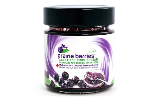 A glass jar with a black lid and a white and purple label. The label contains images of several of the Saskatoon berries.