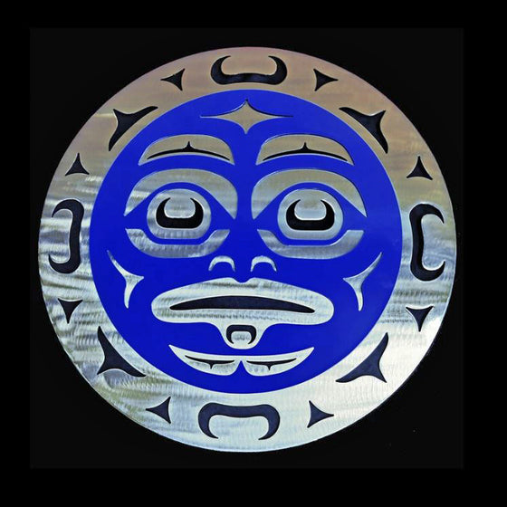 A sculpture of a moon face made by layering carved blue and silver metal. Strategically carved u-shapes and triangles create the illusion of a face with wide eyes and a slightly open mouth.