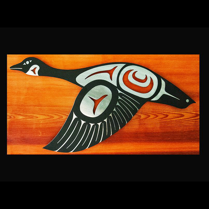 A Canada goose in flight done in Haida style. The brushed steel base provides brilliant contrast to the black powder-coated top layer, creating a dazzling, textured piece of wall art. This goose has its wings down.