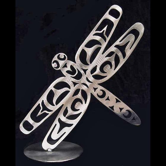 A brushed steel dragonfly done in Haida art style. It is mounted to a sturdy base at one wing, creating the impression that the dragonfly is hovering in midair.