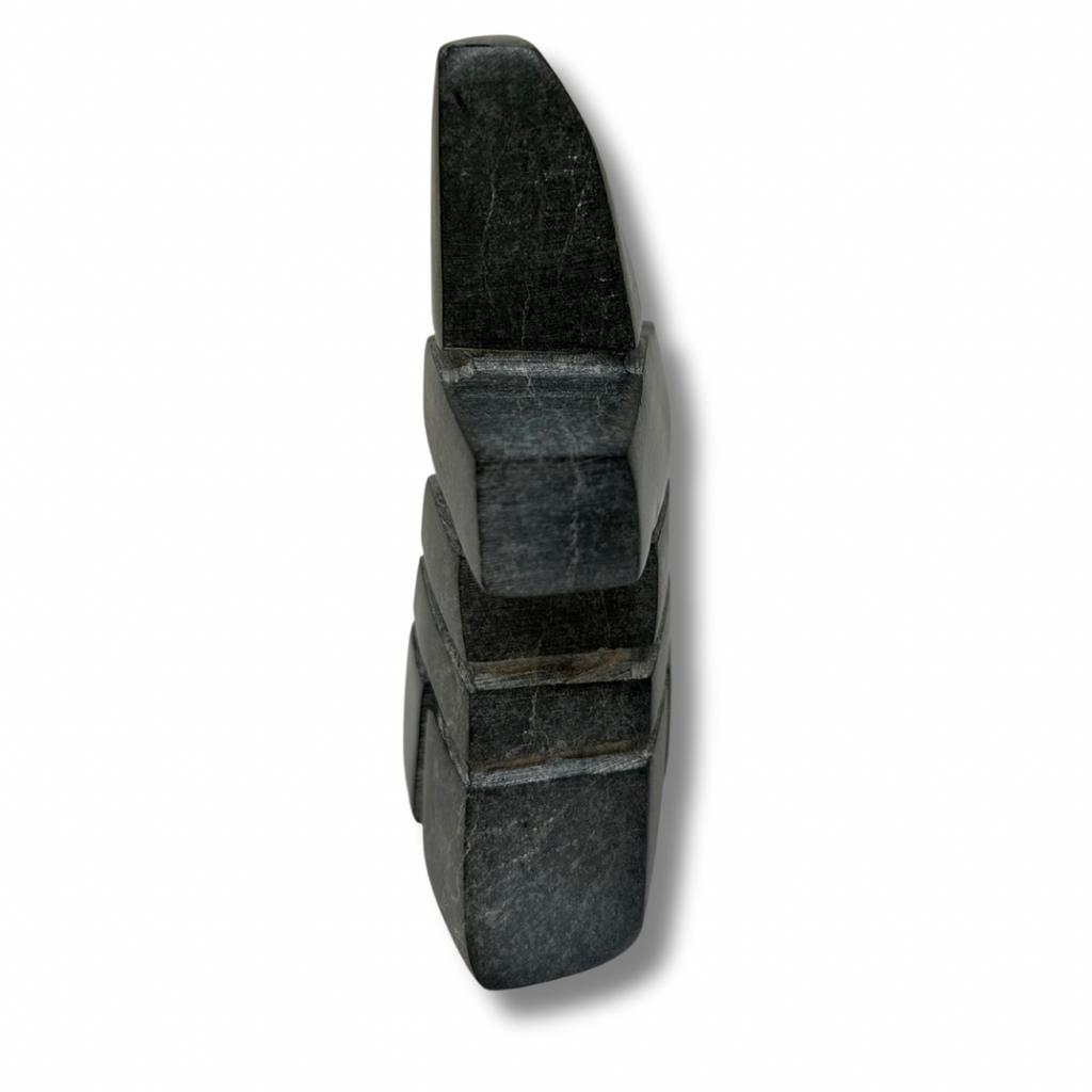 An angular inukshuk in the shape of a person standing in with feet shoulder-width apart. The inukshuk is carved from black stone with stones tightly fitted to each other.