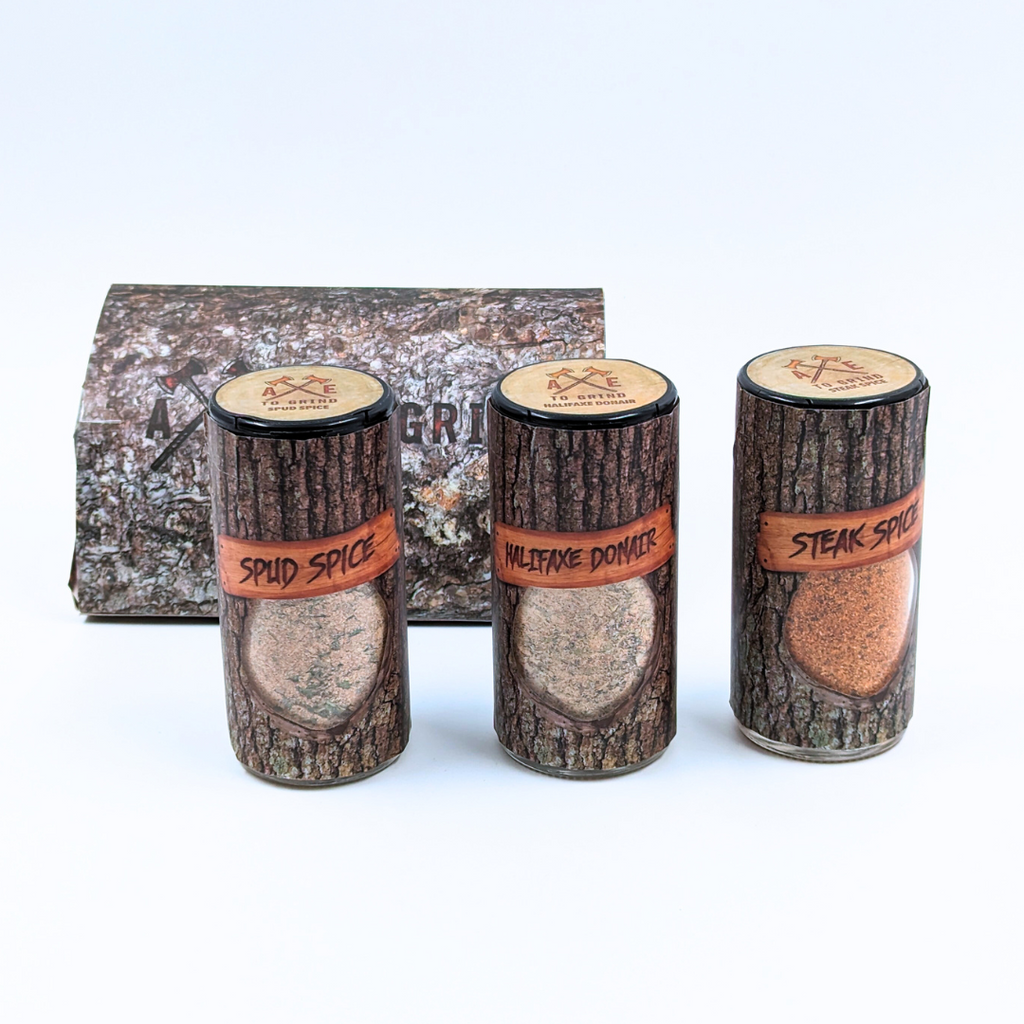 Three separate containers that all have the same packaging. Spice blend is packaged in a reusable/recyclable glass jar printed to look like a log of firewood. The spice mixture is visible through a clear window in the packaging. Larger packaging  with "Axe To Grind" logo on box that looks like firewood.
