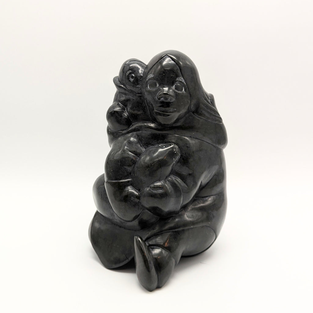 An Inuk woman sits with a child on her back; she holds a seal in her lap. Her left foot is straight out in front of her, the right tucked under her. The sculpture is carved of jet black stone, with expressive faces on each of the three figures. The woman faces the viewer in this picture.