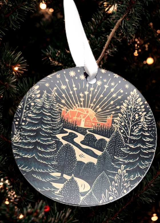 Round Christmas ornament with a winter scene of a house and trees, hanging on a tree.