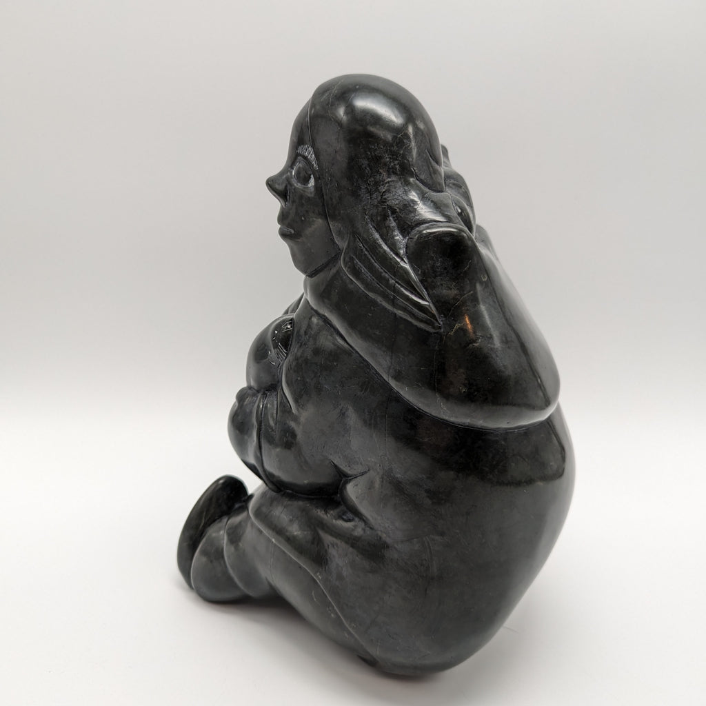 An Inuk woman sits with a child on her back; she holds a seal in her lap. Her left foot is straight out in front of her, the right tucked under her. The sculpture is carved of jet black stone, with expressive faces on each of the three figures. The woman faces away in this picture.