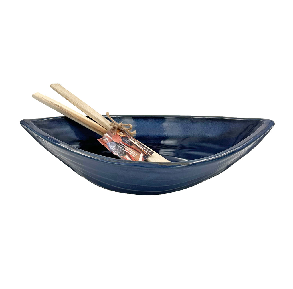 Bottom half of bowl is off-white. Top half of bowl is blue with white accents. Bowl is oval shaped with points at two opposite ends. Two wooden paddles sitting inside the bowl.
