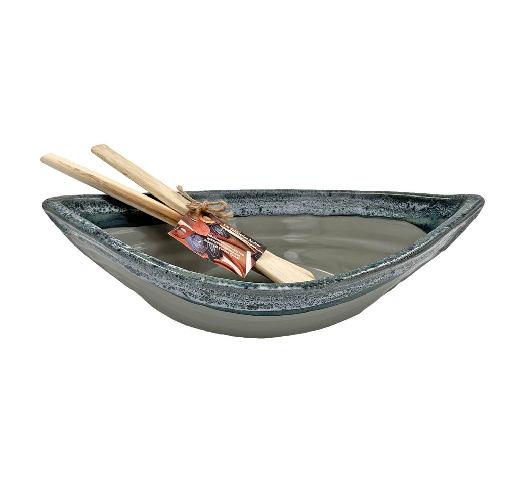 Bottom half of bowl is blue-grey. Top half of bowl is blue with white accents. Bowl is oval shaped with points at two opposite ends. Two wooden paddles sitting in the bowl.