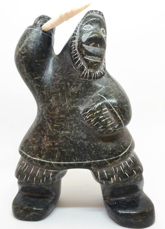 Soapstone carving of Inuit hunter figure. The etchings marked in the stone gives the figure the appearance of wearing a large hooded jacket and boots. Figure has right arm upstretched with hunting tool in hand. The tool is a carved white bone with jagged end.