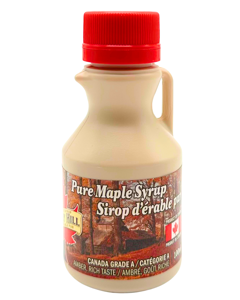 A small cream coloured jug with a picture of a forest going around it. Has the words "Pure Maple Syrup" written on it in both English and French. Has a red cap.
