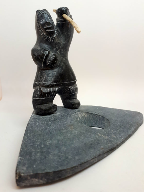 Soapstone carving of Inuit seal hunter figure. The etchings marked in the stone gives the figure the appearance of wearing a large hooded jacket and boots. Figure has left arm upstretched with hunting tool in hand. The tool is a carved white bone with jagged end. Hunter is standing on a stone platform representing frozen water. There is a hole where the hunter waits for the seal to appear.