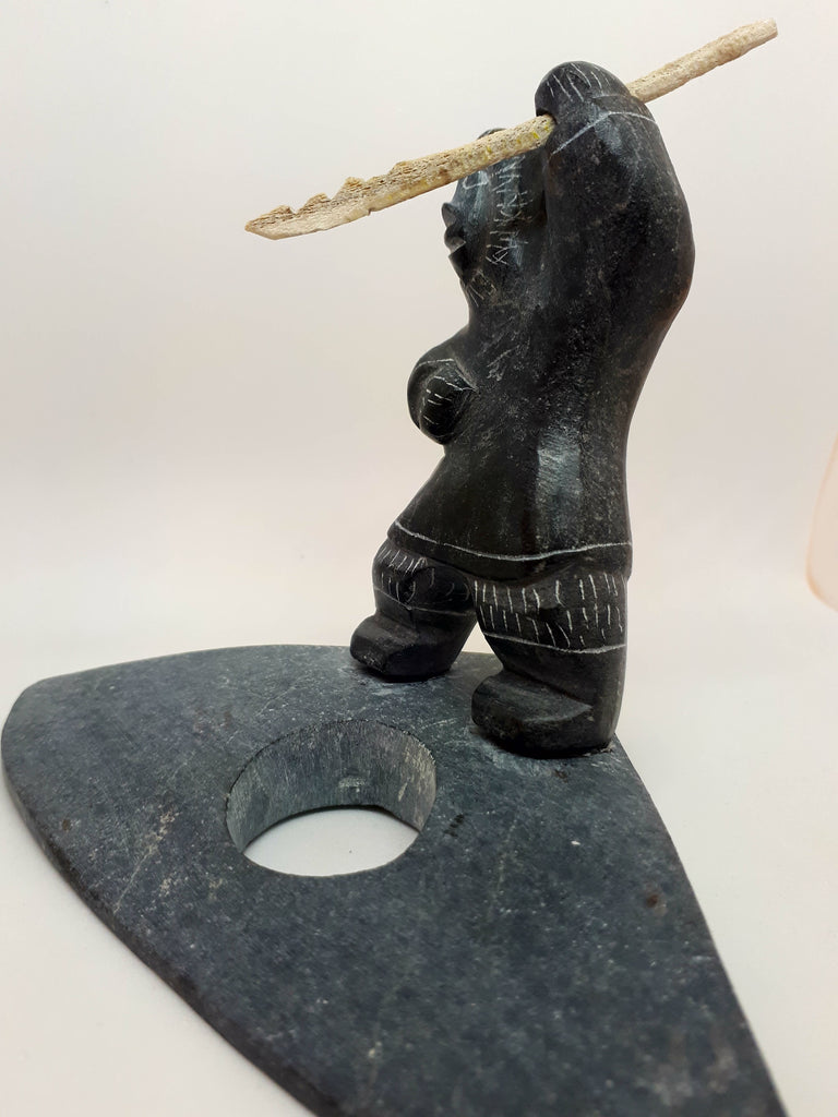 Side view of soapstone carving of Inuit seal hunter figure. The etchings marked in the stone gives the figure the appearance of wearing a large hooded jacket and boots. Figure has left arm upstretched with hunting tool in hand. The tool is a carved white bone with jagged end. Hunter is standing on a stone platform representing frozen water. There is a hole where the hunter waits for the seal to appear.