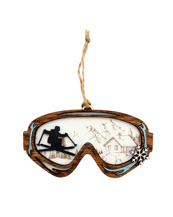 Decorative ornament shaped like goggles with a skier and a lodge in the reflection. There is a small snowflake on the side of the goggles.
