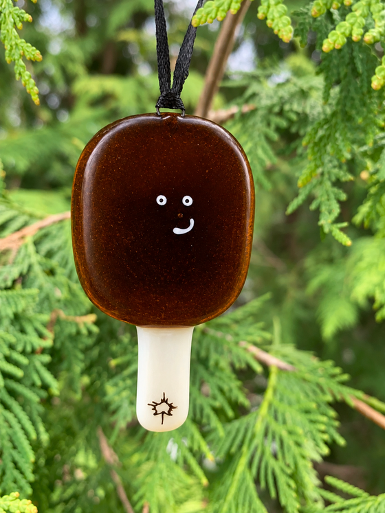 An adorably quirky maple taffy ornament that is Canadian made and made of fused glass. The ornament is dark in colour and round, and the taffy has a cute smiling expression.