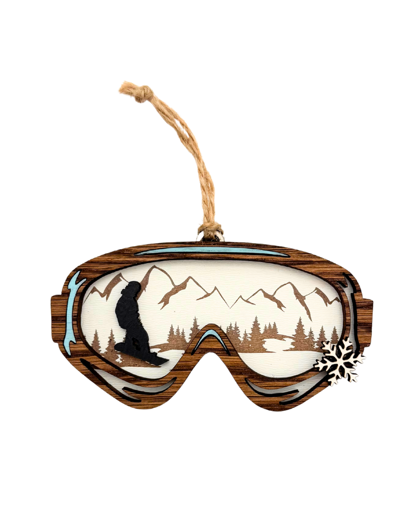 Wooden ornament shaped like ski goggles with a mountains and a snowboarder in the reflection and a small snowflake on the side of the goggles.