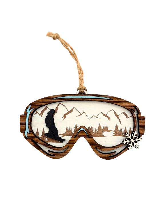 Wooden ornament shaped like ski goggles with a mountains and a snowboarder in the reflection and a small snowflake on the side of the goggles.