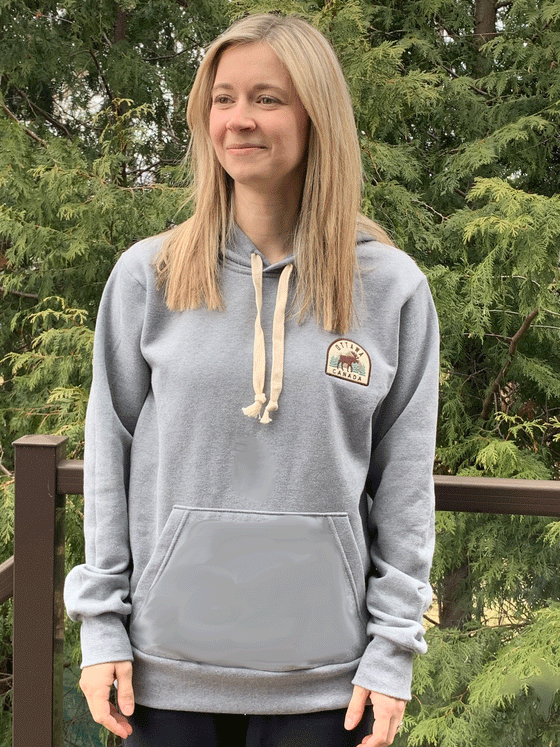 A grey hoodie with white drawstrings. A crest of a brown moose against green trees, grey mountains and a cream background, with Ottawa, Canada written in brown above and below is on the left chest. The crest has a brown outline.