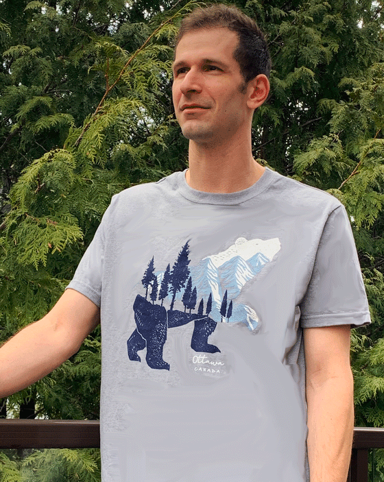 A grey shirt. There is a bear in the middle. The head and neck of the bear are white and light blue mountains. The body and legs of the bear is dark blue trees and the ground. Underneath one of the front bear paws is text that says "Ottawa Canada".