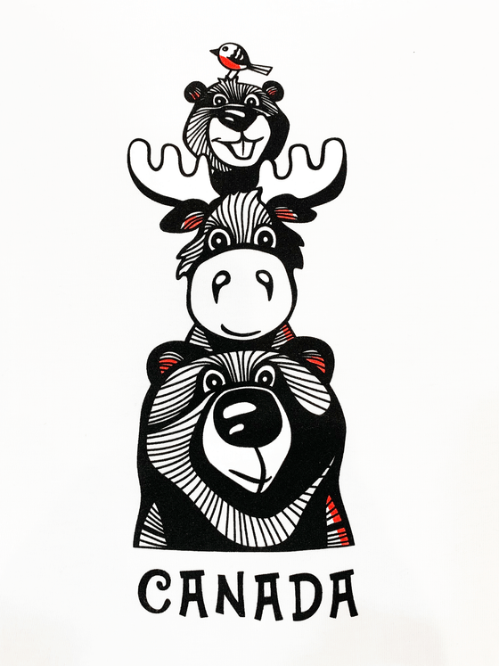 A white shirt. There is a bear, moose, beaver, and a small bird all stacked on each other. The animals are all outlined in black with finer black lines inside. There is red detailing in the ears and the birds stomach. Underneath the bear is written "Canada".