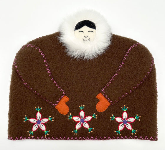 This tea cozy is in the shape of a smiling person wearing a brown parka with white fur around the face. The arms are stitched into the front with blue string. Small mitten covers hand poke out from the sleeves, and three flowers are stitched onto the piece along the bottom.