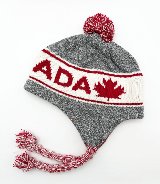 A grey hat with a white band across it. In the white band is written "Canada" and has a maple leaf at the end. There is a red and white pom pom as well as strings.
